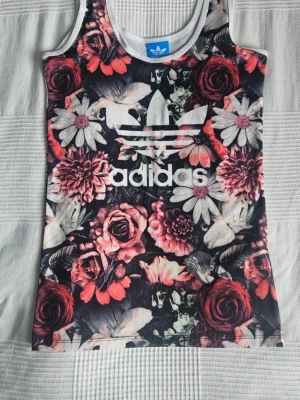 Blommig linnetopp från Adidas - Snygg linnetopp från Adidas med stort vitt logotryck framtill. Toppen har ett färgstarkt blommönster i rött, rosa, vitt och svart. Storlek S. 