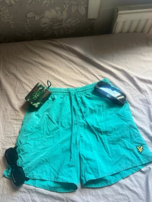  badbyxor    - Snygga turkosa badbyxor med resår och dragsko i midjan. På vänster ben finns en broderad gul fågellogga. Lätta och snabbtorkande, perfekta för stranden eller poolen.Pris kan diskuteras.