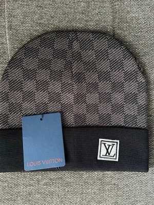 Svart och grå mössa - Snygg svart och grå mössa med klassiskt rutmönster och logga framtill. Mössan har vikt kant och är tillverkad i mjukt stickat material som känns lyxigt och trendigt. Perfekt accessoar för dig som vill ha en exklusiv touch på din outfit.