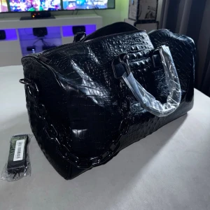 Lyxig Weekendbag / Väska – Svart Krokodilmönster - Säljer en helsvart och lyxig väska i snyggt krokodilmönster som passar lika bra för tjejer som killar. Väskan har en unik svart kedja i plast som gör att den sticker ut, samt ett medföljande långt band för att bära den över axeln. Perfekt som weekendbag, rymlig handväska eller som ett snyggt handbagage på resan. Finns ett fack med dragkedja inuti sidan, lite större än en iphone pro max. Skick: Helt ny. Mått: 46 cm lång, 26 cm hög, 22 cm djup