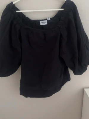Svart topp med puffärmar från Aware - Trendig svart topp från Aware med bred resår vid halsen och stora puffärmar. Toppen har en lös passform och är perfekt för dig som vill ha en stilren men ändå statement look. Tillverkad i ett mjukt tyg som känns skönt mot huden.💕💕💕