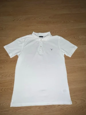 Vit pikétröja från Gant - Klassisk vit pikétröja från Gant med broderad logga på bröstet. Tröjan har korta ärmar, krage och knäppning med tre knappar framtill. Perfekt för en stilren och avslappnad look.