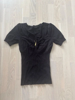 Svart topp med dragkedja - Snygg svart topp med korta ärmar och en liten guldfärgad dragkedja framtill vid urringningen. Toppen har en figurnära passform och är perfekt för att lyfta din outfit med en enkel men cool detalj.