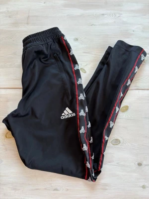 Svarta Adidas träningsbyxor - Snygga svarta träningsbyxor från Adidas med vita loggor längs sidorna och röda detaljer. Byxorna har resår i midjan och två fickor framtill. Perfekta för gymmet eller sportaktiviteter.