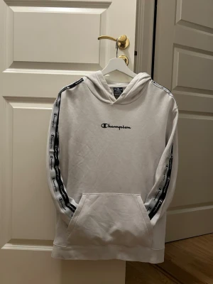 Vit Champion hoodie med logga - Snygg vit hoodie från Champion med svart logga på bröstet och svarta band med Champion-tryck längs ärmarna. Klassisk huva och stor magficka framtill. Tillverkad i mjukt material, perfekt för en avslappnad streetwear-look. Storlek L