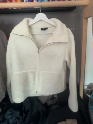 Vit teddyjacka med dragkedja lätt att limma - Mysig vit teddyjacka med dragkedja framtill och stor krage. Jackan har två stora fickor framtill och är gjord i fluffigt, mjukt material som ger en avslappnad look. Perfekt för att hålla värmen under kyliga dagar.