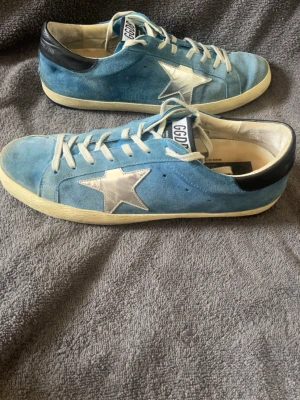 Golden Goose skor - Säljer ett par snygga blå sneakers med vit sula och silverfärgad stjärna på sidan. Skorna har vita snören och svart detalj vid hälen. Ovandelen är i mocka och de har en klassisk låg profil. Perfekta för dig som vill sticka ut lite extra.