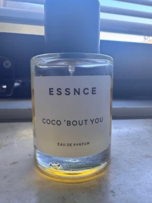 ESSNCE Coco 'Bout You parfym - Minimalistisk parfymflaska i glas med svart lock och vit etikett. Doften heter Coco 'Bout You och är en Eau de Parfum från ESSNCE. Vätskan är gyllengul och flaskan har en stilren, modern design som passar perfekt på sminkbordet.