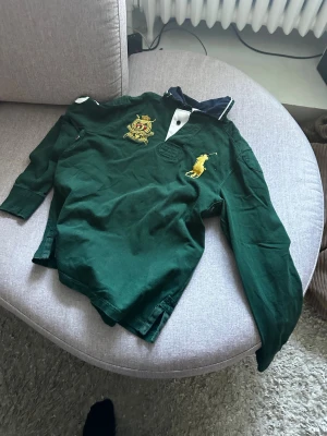 Grön långärmad rugbytröja Polo Ralph Lauren!  - Mörkgrön långärmad rugbytröja från Polo Ralph Lauren med klassisk krage, knappslå och stora broderade loggor i gult och rött på bröstet. Tillverkad i mjuk bomull och perfekt för en sportig och avslappnad stil. 
