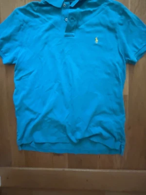 Blå pikétröja med krage - Snygg blå pikétröja med klassisk krage och knappar framtill. Tröjan har korta ärmar och en liten gul broderad logga på bröstet. Perfekt till jeans eller shorts för en avslappnad stil.
