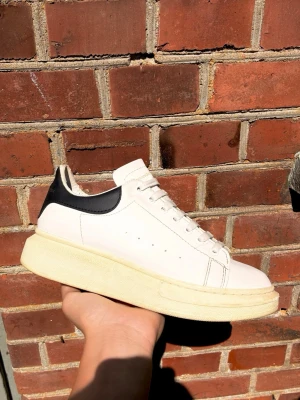 Vita Alexander McQueen sneakers - Stilrena vita sneakers från Alexander McQueen med svart hälparti och tjock sula. Skorna har klassisk låg profil, vita skosnören och diskreta ventilationshål på sidan. Tillverkade i läder med gulddetaljer på hälen.