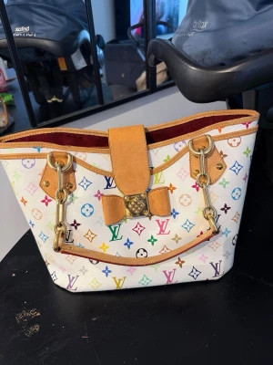 Louis Vuitton multicolor handväska - Säljer en ikonisk Louis Vuitton handväska i vitt med färgglatt monogrammönster. Väskan har detaljer i ljust brunt läder, guldfärgade metalldelar och en unik dekorativ låsdetalj framtill. Perfekt för dig som vill sticka ut med en klassisk designeraccessoar.