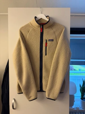 Patagonia Fleecejacka - Säljer en beige teddyjacka från Patagonia med hel dragkedja framtill och hög krage. Jackan har mörkblå detaljer vid dragkedja och ärmslut samt en bröstficka med röd dragkedja. Tillverkad i mjukt, fluffigt material som håller dig varm. Strl M.