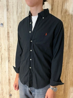 Svart skjorta med knappar - Klassisk svart skjorta med långa ärmar och knappar framtill. Skjortan har en stilren design med liten broderad logga på bröstet och är tillverkad i bomull. Perfekt till jeans eller chinos för en clean look.