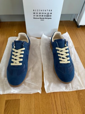 Blå Maison Margiela sneakers - Snygga blå sneakers från Maison Margiela med beige snörning och gummisula. Skorna har en klassisk låg siluett och är tillverkade i en mix av mocka och läder. Perfekta för dig som vill ha en stilren och trendig look.