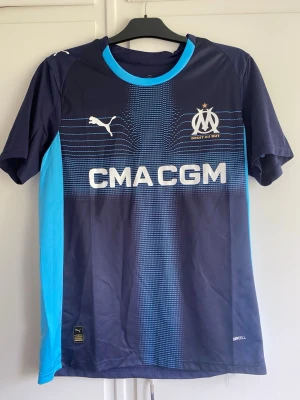 Marseille matchtröja Puma blå - Snygg Marseille matchtröja i mörkblått och ljusblått från Puma. Tröjan har tryck med klubbmärke, sponsorlogga och spelarnamn Greenwood med nummer 10 på ryggen. Tillverkad i lätt och ventilerande material, perfekt för fotbollsträning eller match.