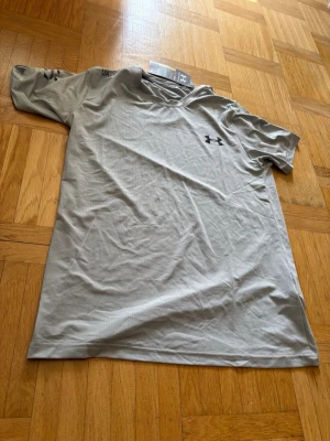 Ljusgrå träningströja från Under Armour - Snygg ljusgrå kortärmad träningströja från Under Armour med svart logga på bröstet. Tillverkad i ett lätt och snabbtorkande funktionsmaterial som passar perfekt för träning och sportaktiviteter.