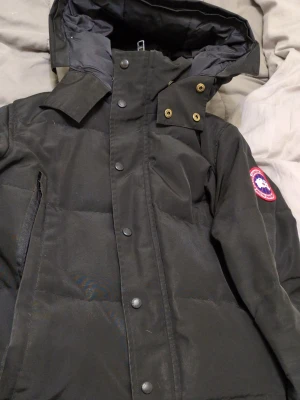 Svart dunjacka från Canada Goose - Säljer en svart dunjacka från Canada Goose med klassisk patch på ärmen. Jackan har hög krage, tryckknappar framtill och diskreta sidofickor. Fylld med dun för extra värme och perfekt för kalla vinterdagar. Enkel, stilren design som funkar till det mesta.