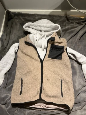 Beige teddyväst med svarta detaljer - Mysig beige teddyväst med dragkedja framtill och svarta detaljer vid fickor och kanter. Västen har två sidofickor och en bröstficka i svart tyg. Perfekt att slänga över en hoodie för en avslappnad look. Bara lägga ett prisförslag. Inga förslag är dumma. (Priset är inte spikat)