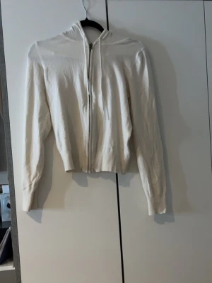 Vit hoodie med dragkedja - Snygg vit hoodie med dragkedja framtill och huva med snören. Modellen är rak med ribbade muddar vid ärmslut och midja. Perfekt för en chill och stilren look. Materialet ser ut att vara mjukt och stickat.