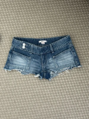 Blå jeansshorts med rå kant - Säljer ett par blå jeansshorts med fransig, rå kant och dubbla knappar i midjan. Shortsen har stora framfickor och bakfickor, samt bälteshällor. Perfekta för varma dagar och en avslappnad stil.