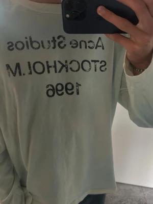 Vit/blå långärmad t-shirt Acne Studios - Vit/blå långärmad t-shirt från Acne Studios med tryck på bröstet där det står 'Acne Studios STOCKHOLM 1996'. Skön passform och enkel design som funkar till det mesta. Perfekt för dig som gillar stilrena plagg med en twist.