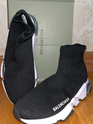 Balenciaga Speed Trainer svarta sneakers - Svarta Balenciaga Speed Trainer sneakers med stickad ovandel i stretchigt tyg och vit chunky sula. Modellen har en strumpliknande passform och tydlig Balenciaga-logga på sidan. Sulan är i gummi med transparent detalj vid hälen för en futuristisk vibe.