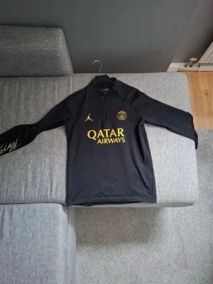Svart PSG träningströja Jordan - Snygg svart långärmad träningströja från PSG x Jordan med gul logga och Qatar Airways-tryck på bröstet. Slim fit-modell med halv dragkedja framtill och detaljer i gult. Perfekt för fotbollsträning eller chill.