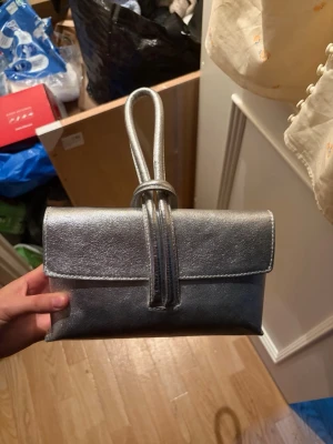 Silvrig kuvertväska med handrem - Snygg kuvertväska i metallic-silver med unik handrem som går över väskans framsida. Väskan har en stilren, rektangulär form och är tillverkad i ett glansigt material som ger en modern look. Perfekt för att lyfta din outfit med en cool detalj 💕😍 aldrig andvänt! Men hoppas någon annan får bättre/mer andvändning för den 🥰kan förhandla om priset men köpt för 1000 kr 