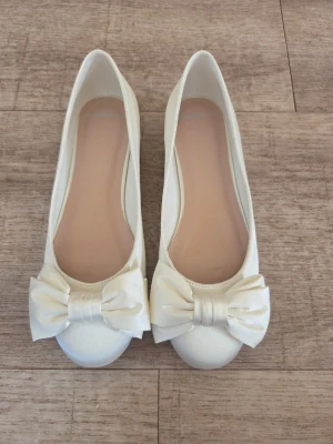 Satinskor i ballerinamodell med rosett st 38 - Snygga vita ballerinaskor i satinliknande material med stora dekorativa rosetter framtill. Skorna har rund tå och låg klack. Färg Ivory i lyxig satin 
