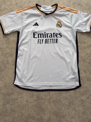 Real Madrid matchtröja Adidas - Snygg vit Real Madrid matchtröja från Adidas med svarta och guldiga detaljer. Tröjan har klubbmärke och Adidas-logga på bröstet samt texten 'Emirates Fly Better' framtill. Tillverkad i lätt och ventilerande material, perfekt för fotbollsträning eller match.
