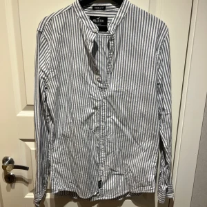 Randig henley-skjorta från Hollister - Snygg randig henley-skjorta från Hollister i vitt och grått. Skjortan har muscle fit-passform, lång ärm och knappar framtill. Tillverkad i stretchig bomullsmix för extra komfort. Perfekt för dig som gillar en clean och avslappnad stil.