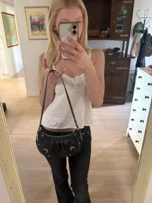 Svart axelremsväska i skinnlook - Snygg svart handväska med axelrem och silverfärgade detaljer. Väskan har rundad form och dekorativa veck framtill. Perfekt storlek för det viktigaste och ger en trendig touch till din outfit. Axelremmen går att göra kortare eller längre. 