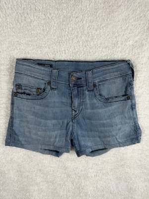 True Religion Geno No Flap Jeansshorts storlek 28 Ljus denim - Snygga blå jeansshorts med klassisk femficksdesign, bälteshällor och knappstängning. Shortsen har en normal passform och är perfekta för varma dagar. De har en ljus tvätt och subtila slitningar för en avslappnad look.