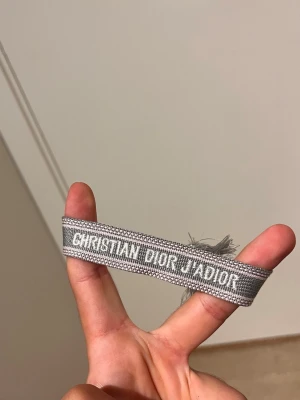 Grått armband Christian Dior - Snyggt grått tygarmband från Christian Dior med texten 'J'ADIOR' broderad i vitt. Armbandet har en enkel och trendig design med fransar i ändarna. Perfekt accessoar för att lyfta din outfit.