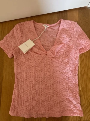 Rosa spetsblus med v-ringning - Supersöt rosa blus i spets med v-ringning och korta ärmar. Blusen har ett fint knut-detalj framtill och är tillverkad i ett lätt och luftigt material. Perfekt för dig som vill ha en feminin och trendig topp till sommaren. Orginalpris 400kr
