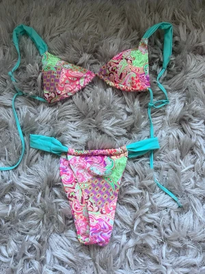 Färgglad bikini med paisleymönster - Säljer en superfin bikini med färgstarkt paisleymönster i rosa, grönt och gult. Både överdel och underdel har turkosa knytband som gör att du kan justera passformen. Perfekt för lata dagar på stranden eller poolhäng med vänner!