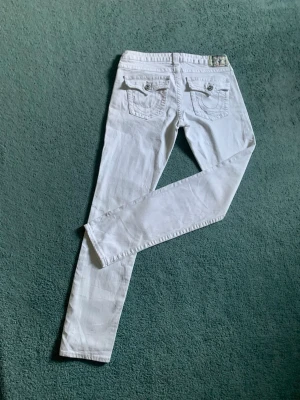 Vita jeans från True Religion - Säljer ett par vita jeans från True Religion med snygga detaljer på bakfickorna och silverfärgade knappar. Jeansen har klassisk femficksdesign och är tillverkade i ett mjukt denimtyg. Perfekta för dig som vill sticka ut med en fräsch look.framsidans fickor är inte riktiga med det är bakfikornas och den lilla fickan på framsidan.pris kan diskuteras 