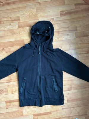 Svart CP Company hoodie med glasögon  - Cool svart hoodie med dragkedja framtill och två fickor med dragkedja. Huvan har inbyggda glasögon . Perfekt för dig som vill sticka ut med en unik detalj. Tillverkad i mjukt material som känns skönt mot huden. Knappt använd tvättad max 5 gånger Äkta. Vid frågor skriv 😊