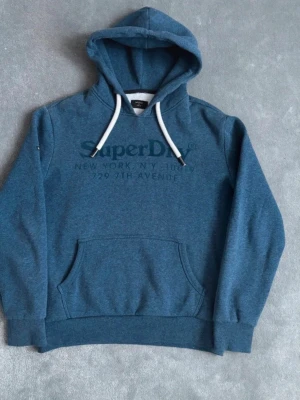 Blå hoodie från Superdry - Säljer en blå hoodie från Superdry med tryck på bröstet och texten 'New York, NY 10019, 729 7th Avenue'. Hoodien har vit dragsko i huvan och en stor magficka. Perfekt för dig som gillar en avslappnad och stilren look.