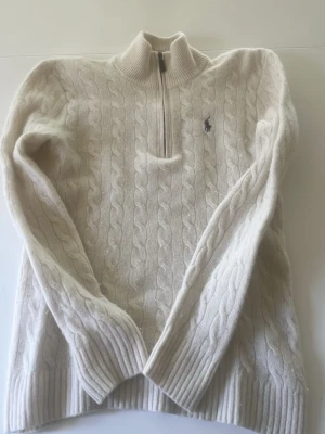 Vit kabelstickad tröja Polo Ralph Lauren - Kabelstickad vit tröja från Polo Ralph Lauren med halv dragkedja och hög krage. Tröjan har ribbade muddar och nederkant samt den klassiska broderade loggan på bröstet. Perfekt för lager på lager och riktigt snygg till jeans. Säljer pga att den blivit för liten, Scick 8/10 då det finns lite tecken på användning, hör av er vid funderingar