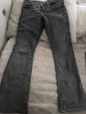 Grå bootcut jeans med fickor från Ginatricot - Säljer ett par grå bootcut jeans i denim med klassisk femficksdesign och knappar på bakfickorna. Jeansen har normal midja och lätt utsvängda ben, vilket ger en avslappnad och trendig siluett.