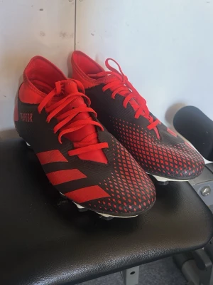 Adidas Predator svart/röda fotbollsskor - Säljer ett par Adidas Predator fotbollsskor i svart och rött med klassiska tre ränder på sidan. Skorna har snörning, mönstrad ovandel och fasta dobbar för bra grepp på planen. Perfekta för dig som vill ha stil och kontroll på fotbollsplanen.
