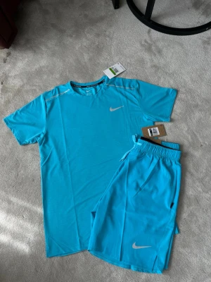 Nike blått träningsset t-shirt & shorts - Snyggt träningsset från Nike i klarblå färg. Setet består av en kortärmad t-shirt och matchande shorts, båda med Nike-logga i vitt. Materialet är lätt och andas, perfekt för träning eller sportaktiviteter. Shortsen har resår i midjan och dragsko för bästa passform.