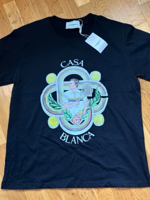 Svart Casa Blanca t-shirt med tryck - Snygg svart t-shirt från Casablanca med stort färgglatt tryck framtill som visar en tennisspelare, tennisbollar och grafiska detaljer. T-shirten har rund hals och korta ärmar. Perfekt för dig som gillar sportig och unik stil.