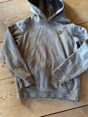 Grå hoodie med gul logga - Säljer en grå hoodie med huva och magficka. På bröstet sitter en broderad gul logga i form av en fågel. Hoodien har ribbade muddar vid ärmar och nederkant, samt en klassisk avslappnad passform. Perfekt för en chill och stilren look.