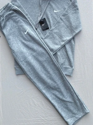 Grått Nike set med hoodie och byxor - Säljer ett grått set från Nike med mjuk hoodie och matchande byxor. Båda plaggen har Nike-logga i vitt och är tillverkade i mjukt sweatshirtmaterial. Hoodien har dragsko och ficka framtill.