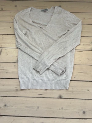 Ljusgrå v-ringad tröja - En stilren ljusgrå tröja med v-ringning och långa ärmar. Tröjan har ribbade muddar vid ärmslut och nederkant, vilket ger en snygg passform. Perfekt att matcha med jeans eller kjol för en enkel och clean look.