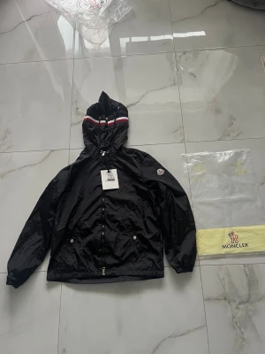 Svart vindjacka från Moncler - Snygg svart vindjacka från Moncler med huva och dragkedja framtill. Jackan har vita och röda ränder på huvan, två fickor med tryckknappar och justerbar dragsko. Tillverkad i glansigt, lätt material. Perfekt för blåsiga dagar.