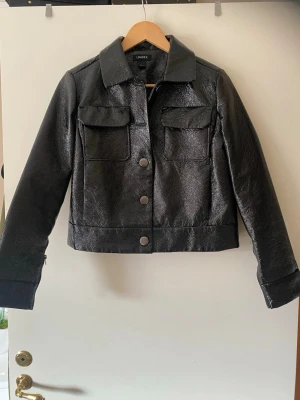 Svart skinnjacka från Lindex - Snygg svart skinnjacka ( polyester)från Lindex med klassisk krage, två bröstfickor med lock och knappar framtill. Jackan har en rak, kortare passform och långa ärmar. Perfekt för dig som vill ha en stilren och trendig look.